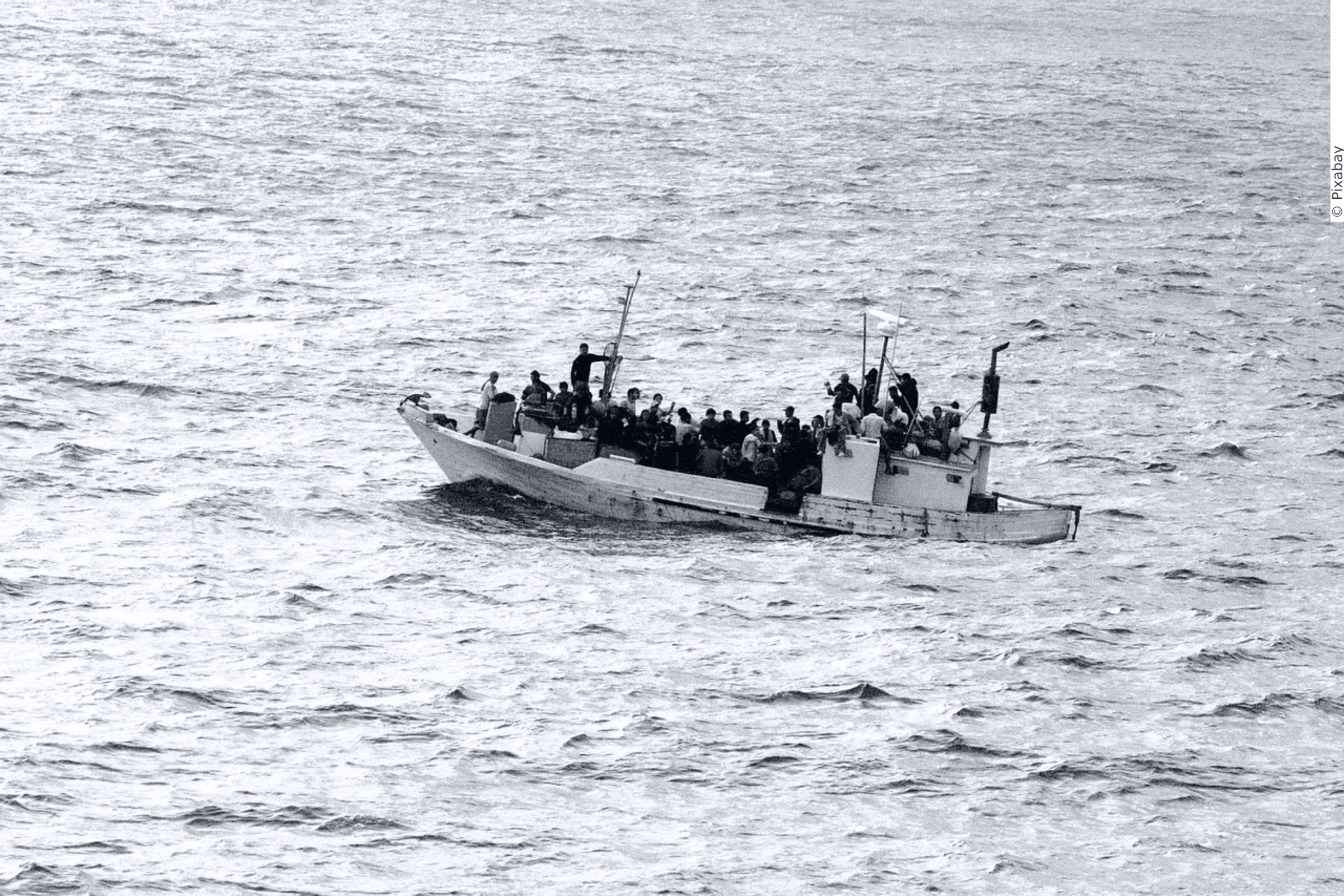 A small boat floats in a vast see. Over 20 people sit or stand in the boat.