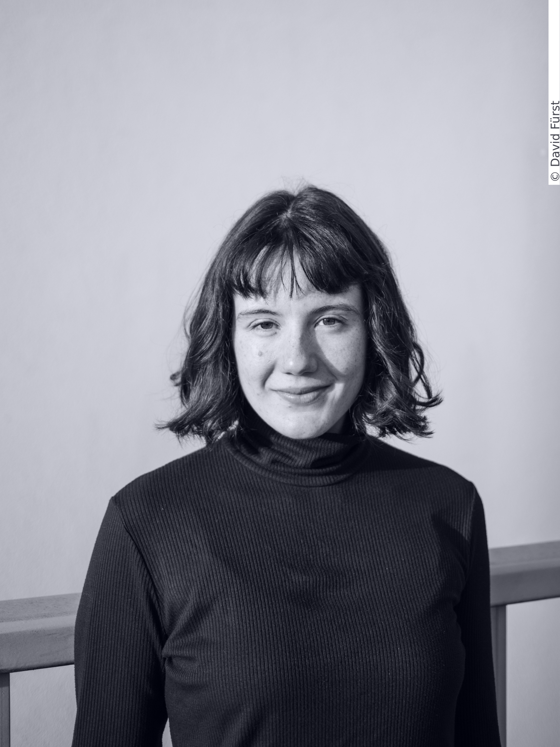 Portrait of Nina Peier. Ms Peier has shoulder-length dark hair and a fringe, dark eyes and freckles. She wears a black turtleneck.