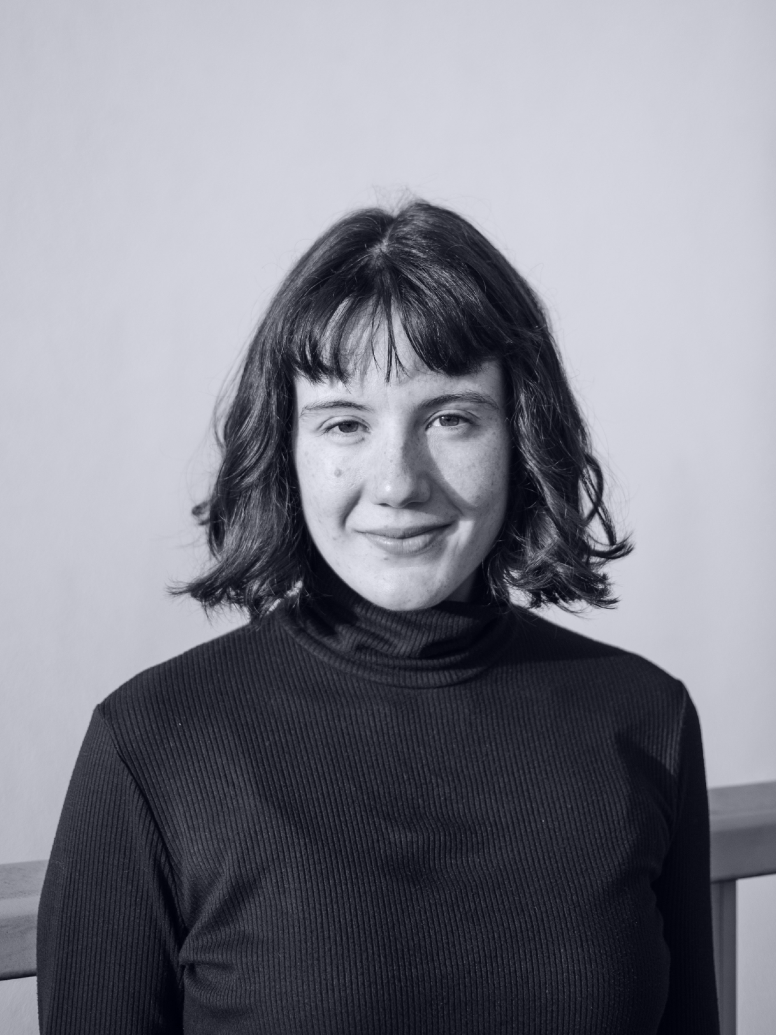 Portrait of Nina Peier. Ms Peier has shoulder-length dark hair and a fringe, dark eyes and freckles. She wears a black turtleneck.