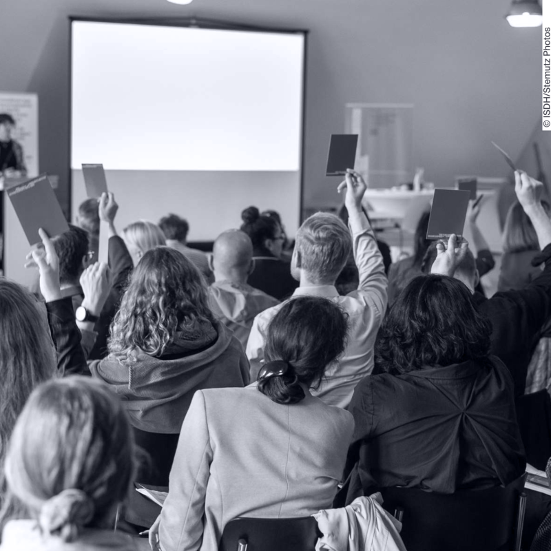 A roomful of people is photographed from behind. They are raising a cardboard card in order to vote.