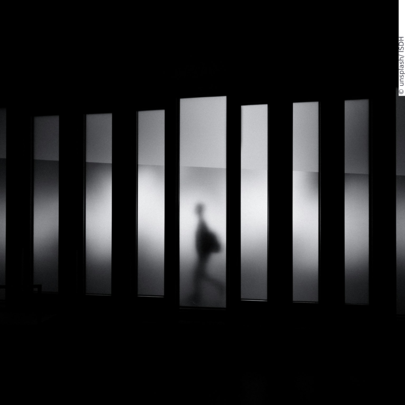 A person walks through light filters that are milky white and occasionally make the person visible. The person is only visible as a walking shadow since the lighting is indirect.