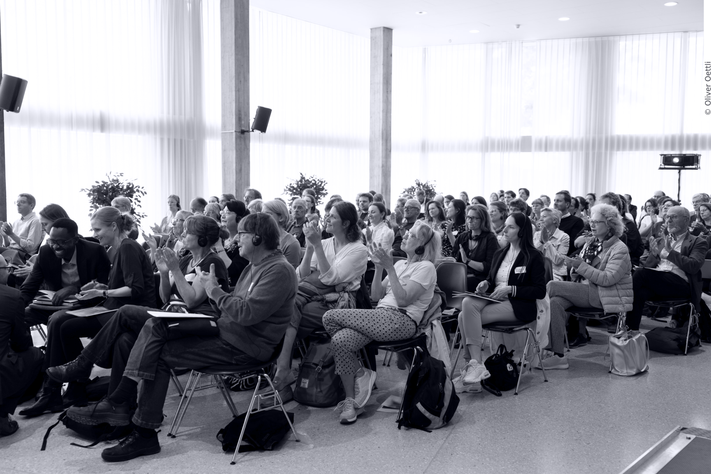 Publikum klatscht in Konferenzraum und hat alle blicken in die gleiche Richtung. Der Saal ist schlicht eingerichtet und mit dünnen Vorhängen bestückt.